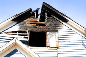 A fire-damaged house in Tacoma, Washington, with a burned roof and smoke-stained blue siding under a clear sky, showing visible structural damage from a recent fire.